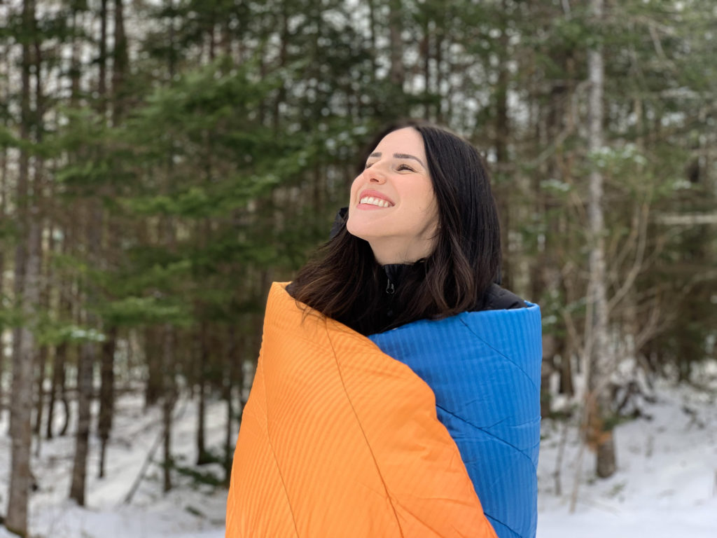 Julia cozied up in the Rumpl Original Puffy Blanket with the snowy forest in the background.
