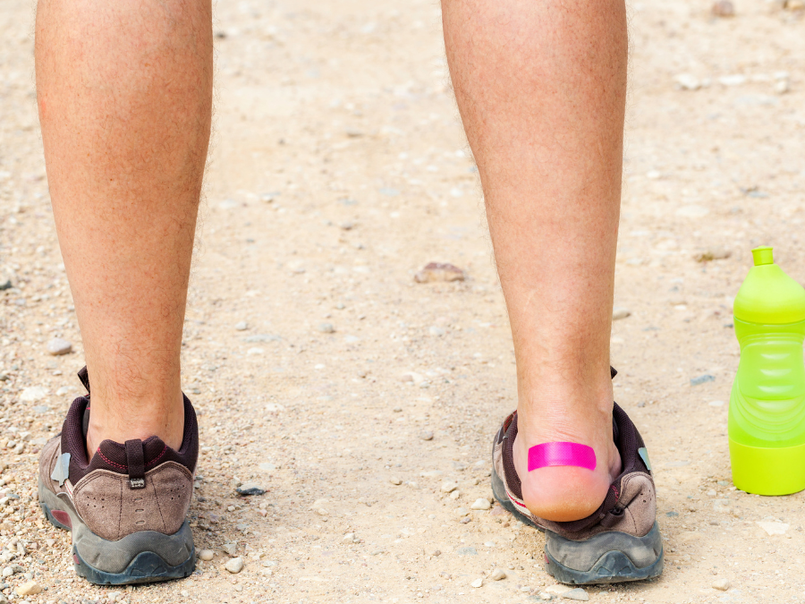 The back of a hikers legs with one hiking shoe off and a pink bandaid over their blisters. 